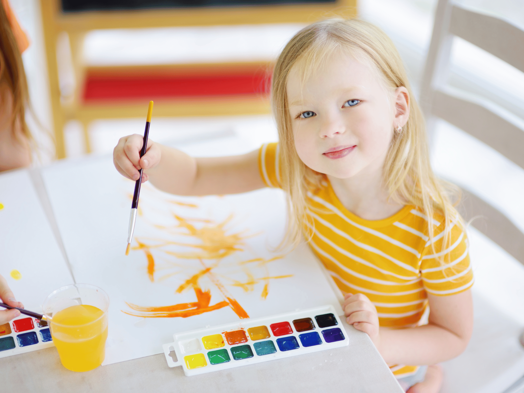 Young girl in a yellow striped shirt paints with a brush, using a watercolor palette and orange paint on white paper at a bright table
